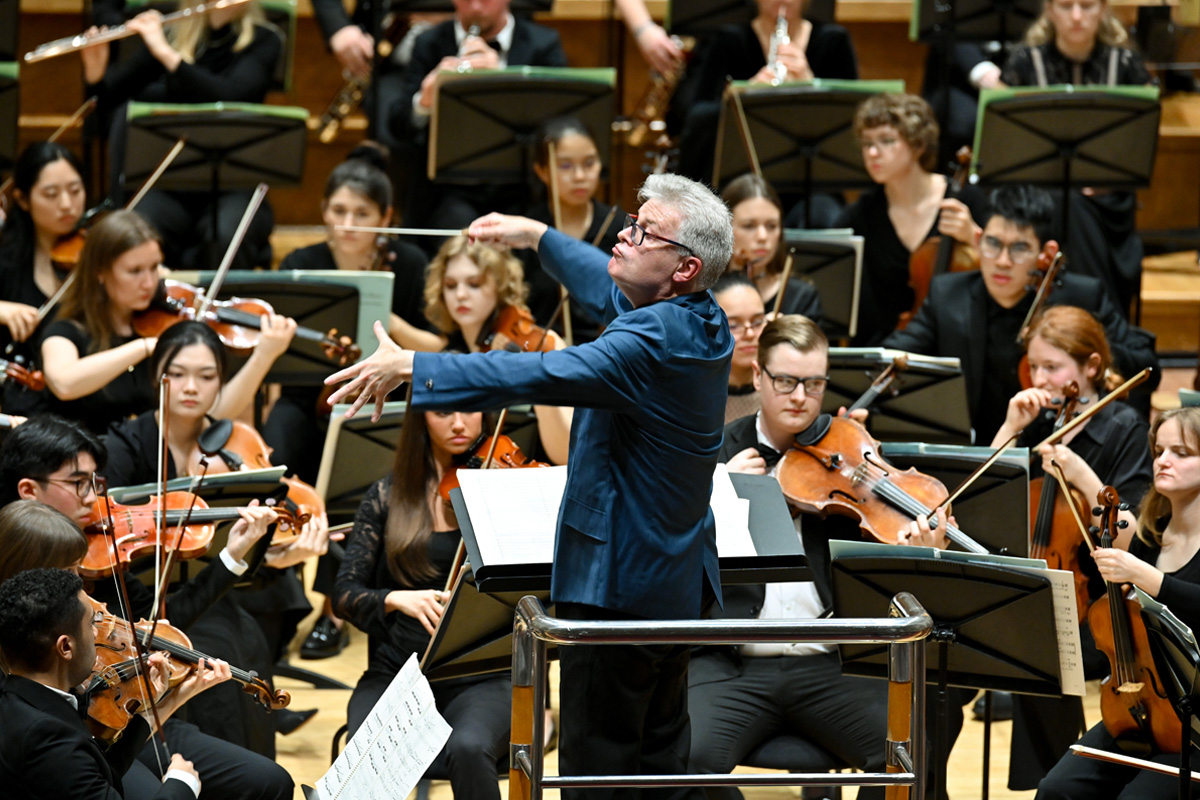 A man wearing a blue suit, leading an orchestra performance with a conductors baton.